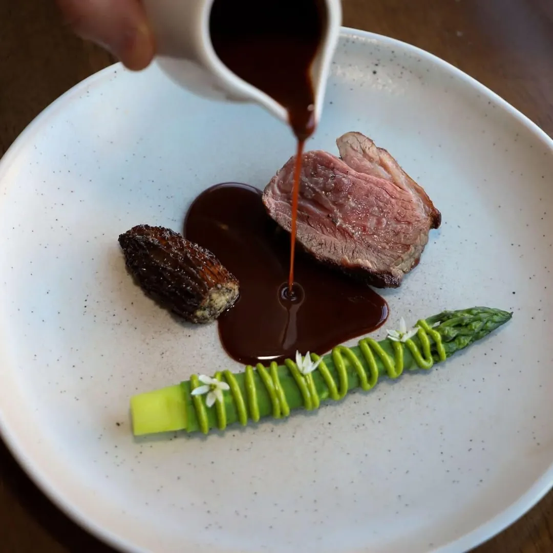 Sauce being poured over sliced meat, with asparagus and a morel